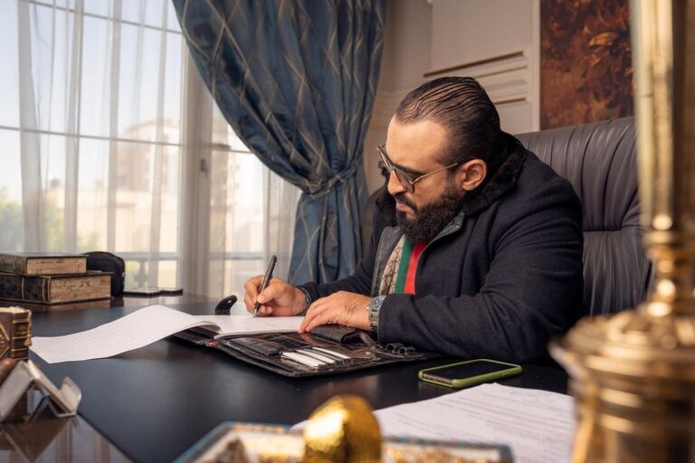 رجل أعمال ملتح بسترة داكنة يوقّع أوراقًا على مكتب خشبي