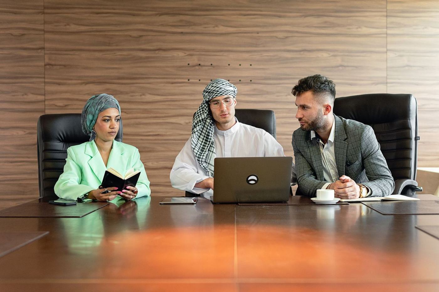 Three colleagues, one in a white kandura and checked ghutra, seated at a boardroom table with a laptop