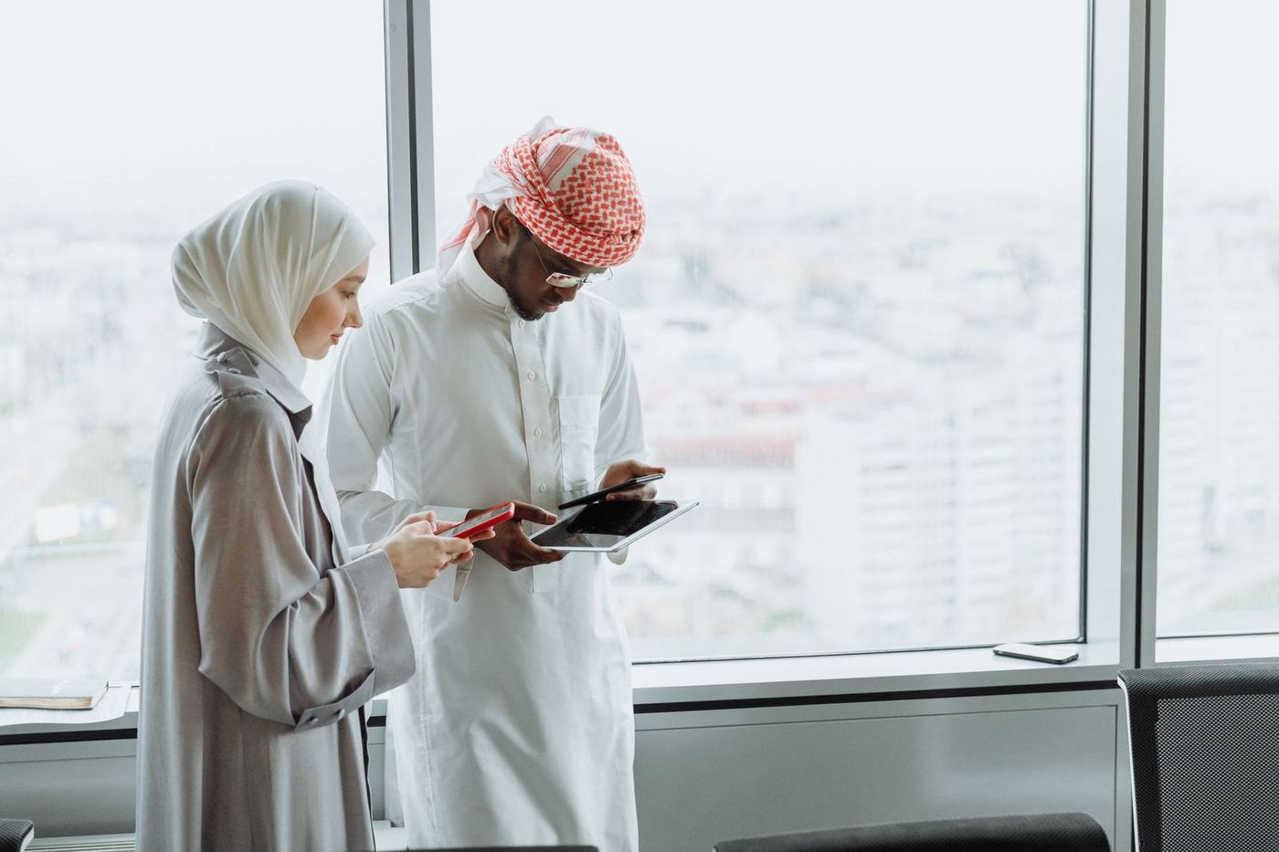 Woman in a white hijab and a man in a kandura and red ghutra reviewing a tablet by an office window
