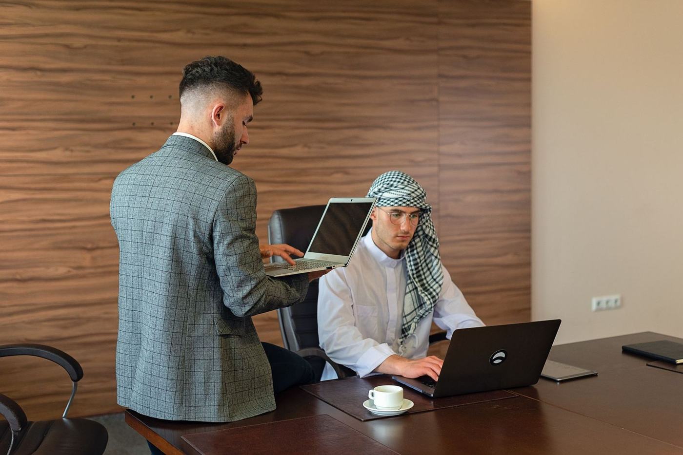 Man in a white kandura and checked ghutra working at a laptop while a colleague in a suit stands beside him