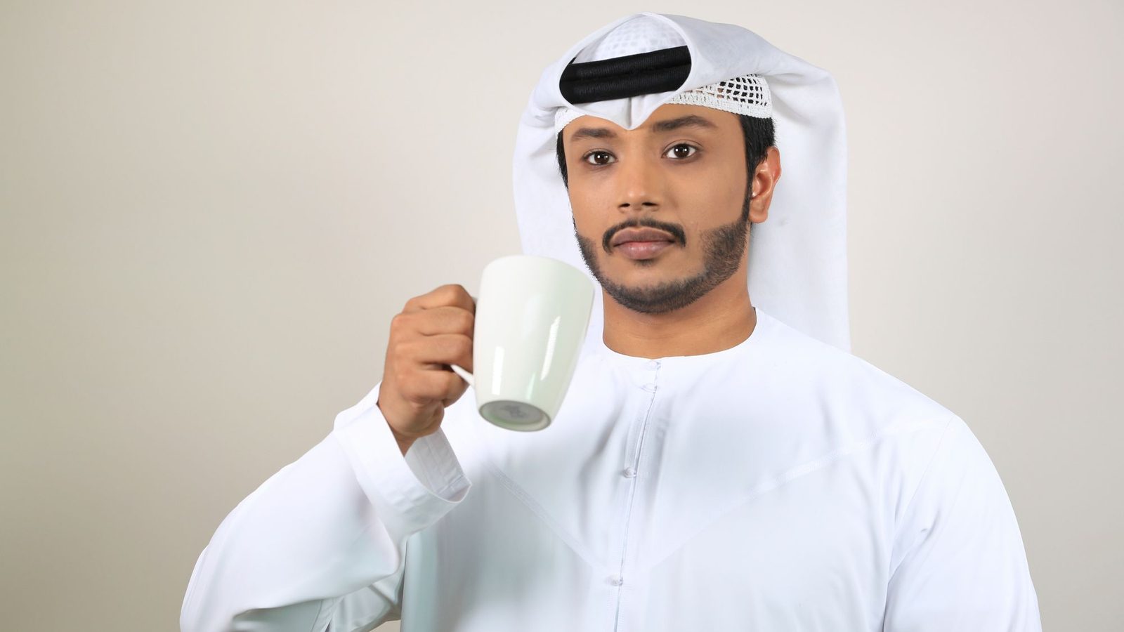 Man in a white kandura and ghutra holding a cup indoors