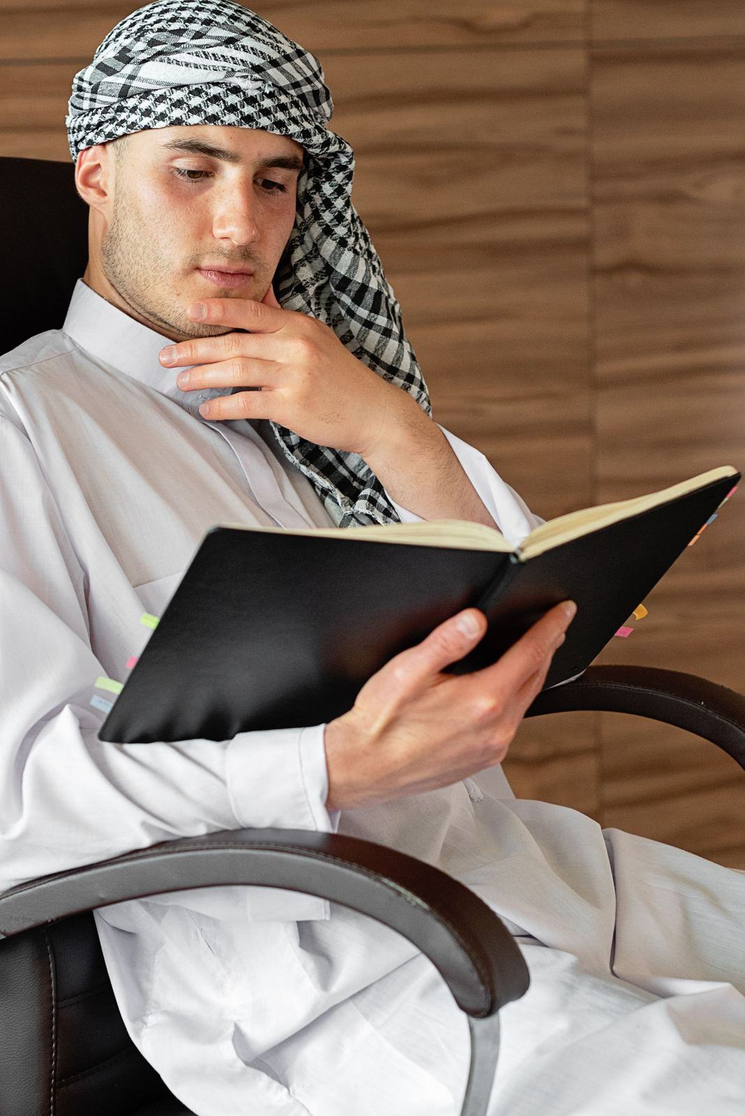 Man in a white kandura and checked ghutra reading a black folder in an office chair