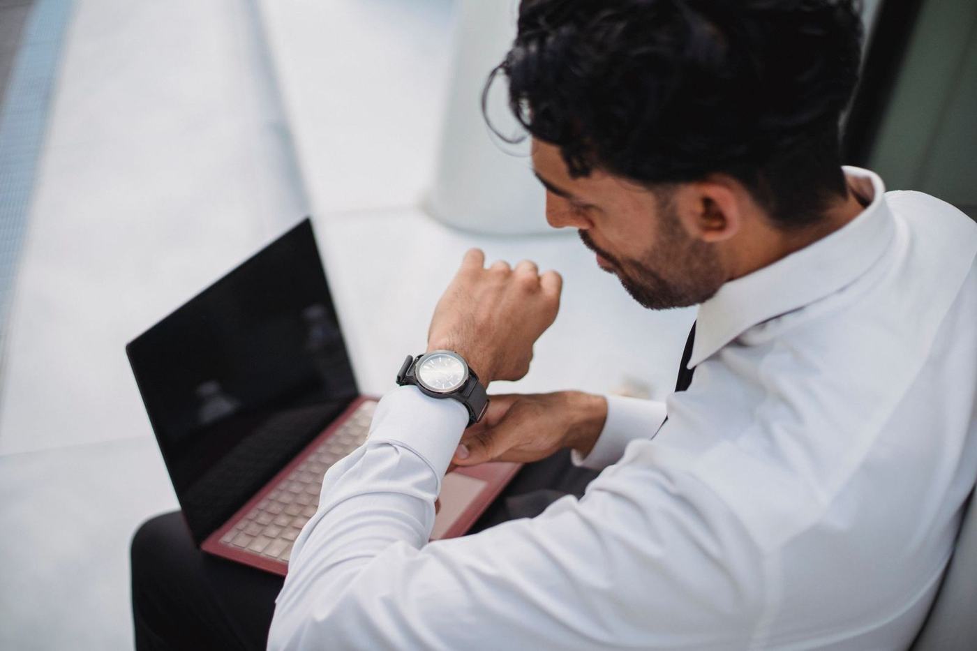 Man in a white shirt checking his watch while working at a laptop