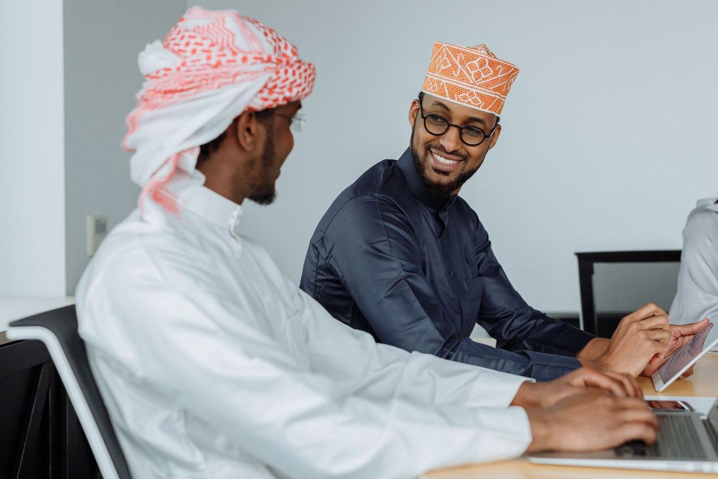 Two men in kandura and traditional caps talking while working on laptops