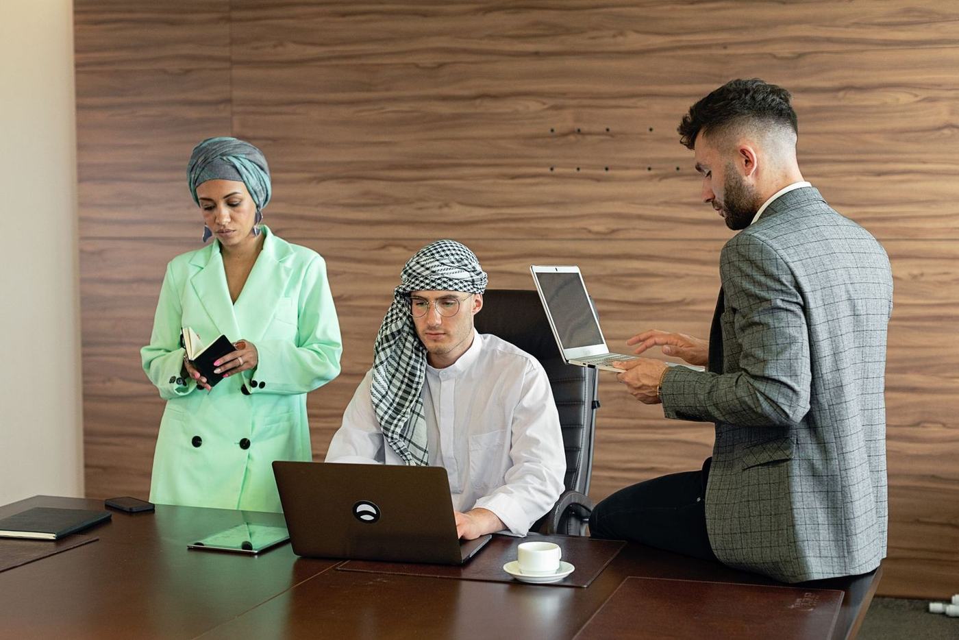Three colleagues at a boardroom desk, one in a kandura and ghutra seated at a laptop