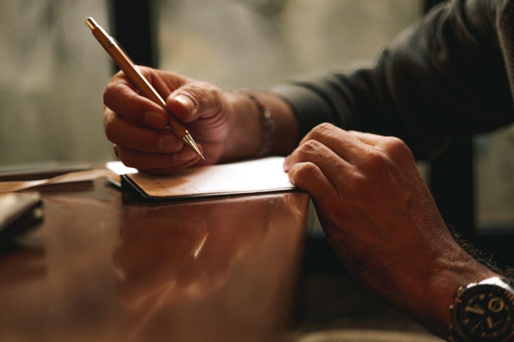 Man writing with a gold pen on a paper at a wooden desk