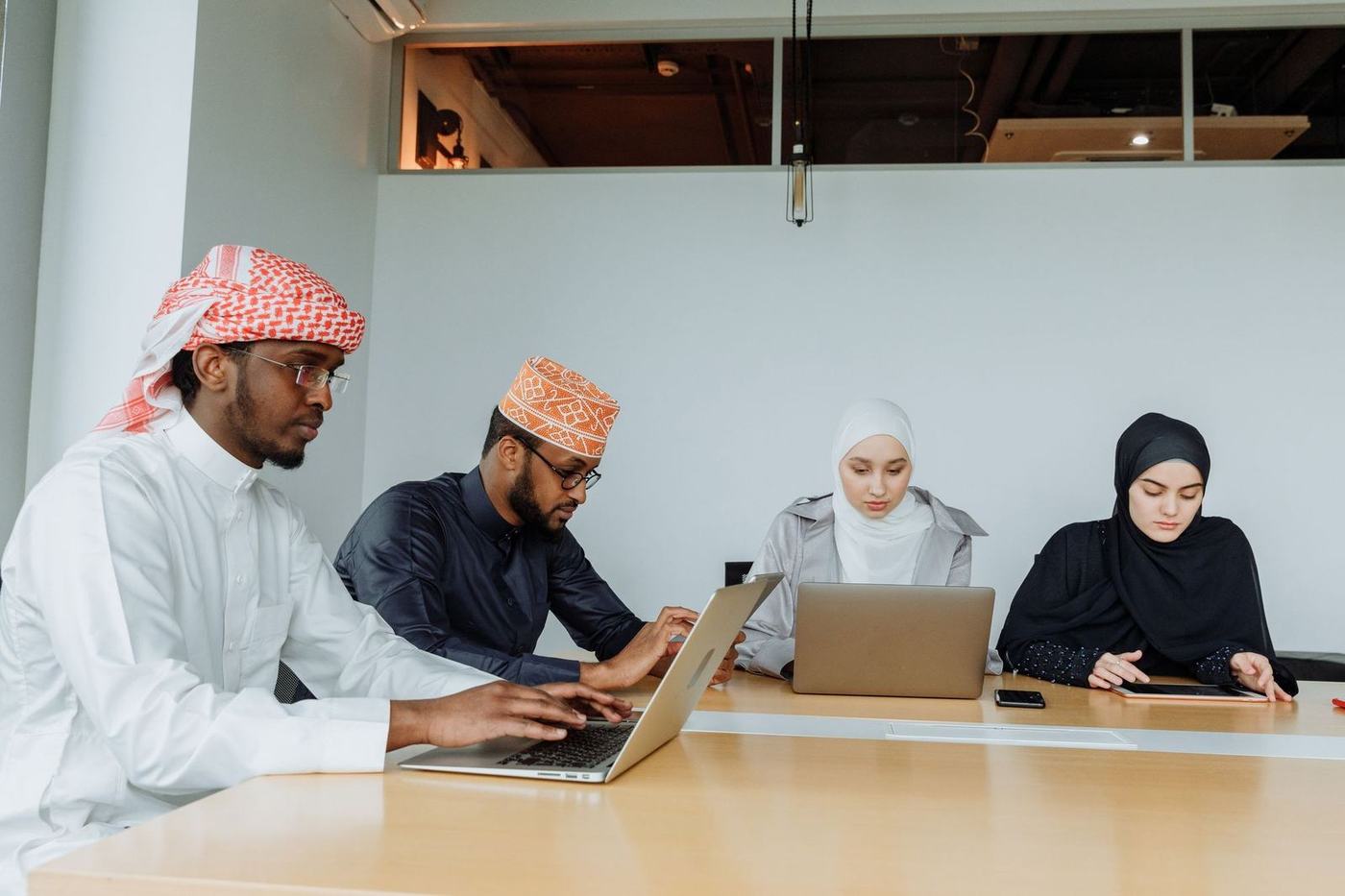 Two men in kandura and two women in hijab working on laptops at a meeting table