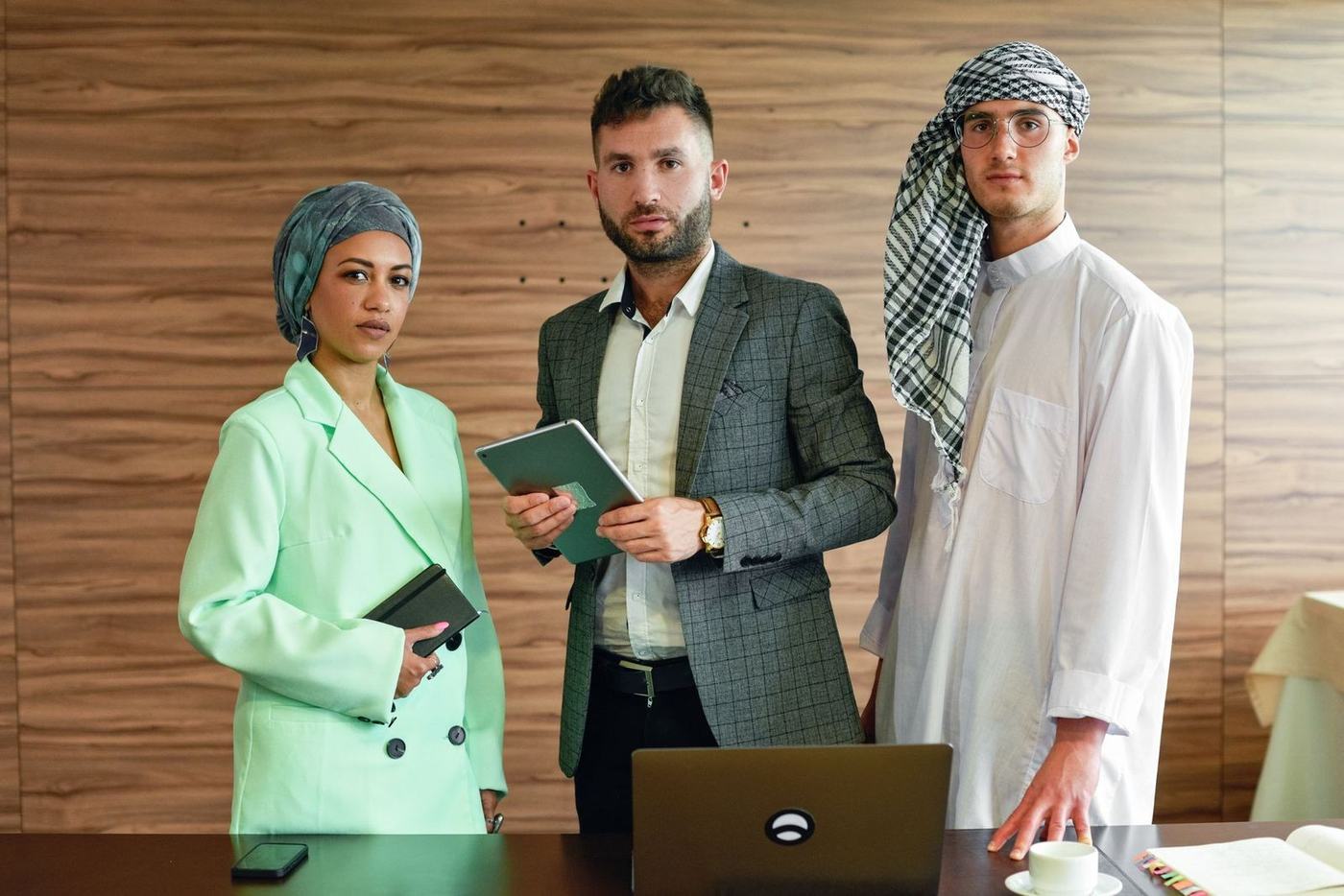 Three business partners, one in a white kandura and ghutra, standing at a boardroom table
