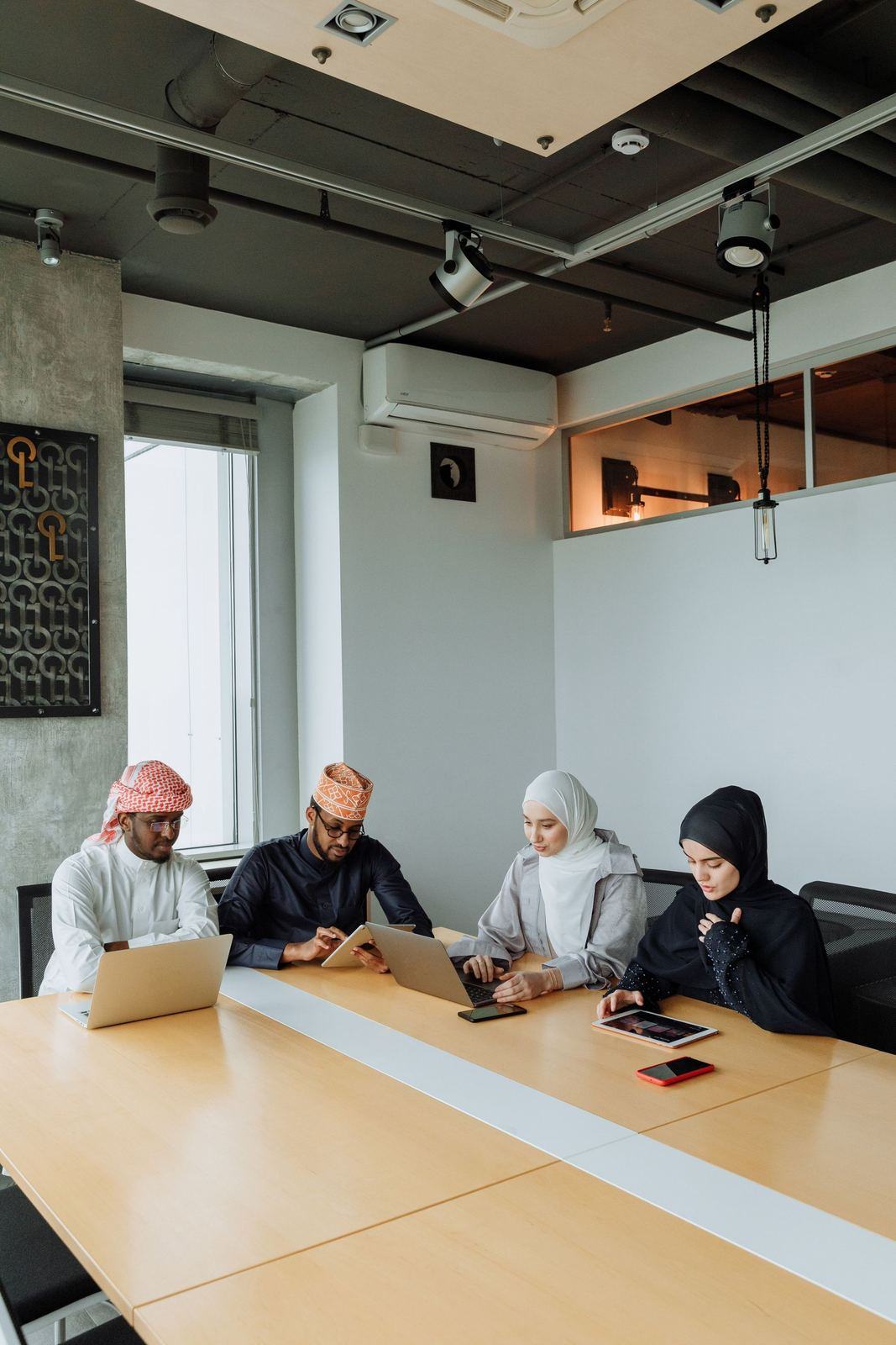 Four colleagues in kandura and hijab around a long meeting table with laptops