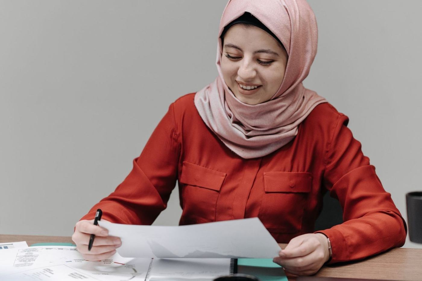 Smiling woman in a pink hijab signing a sheet of paper at a desk