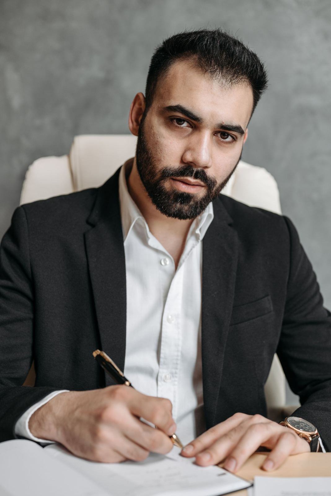 Bearded man in a black suit signing a document at a desk