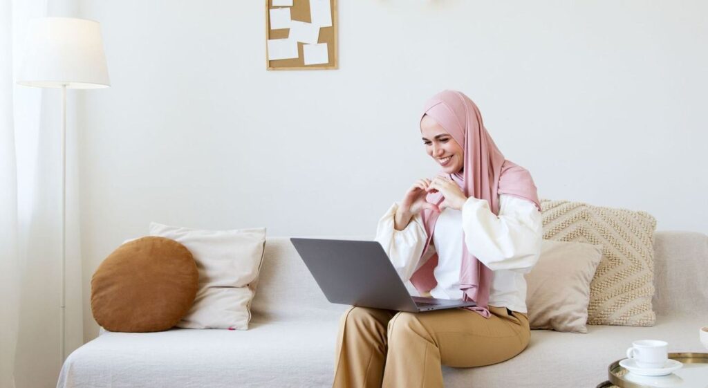 A woman in a hijab on a video call on her laptop, sitting on a sofa