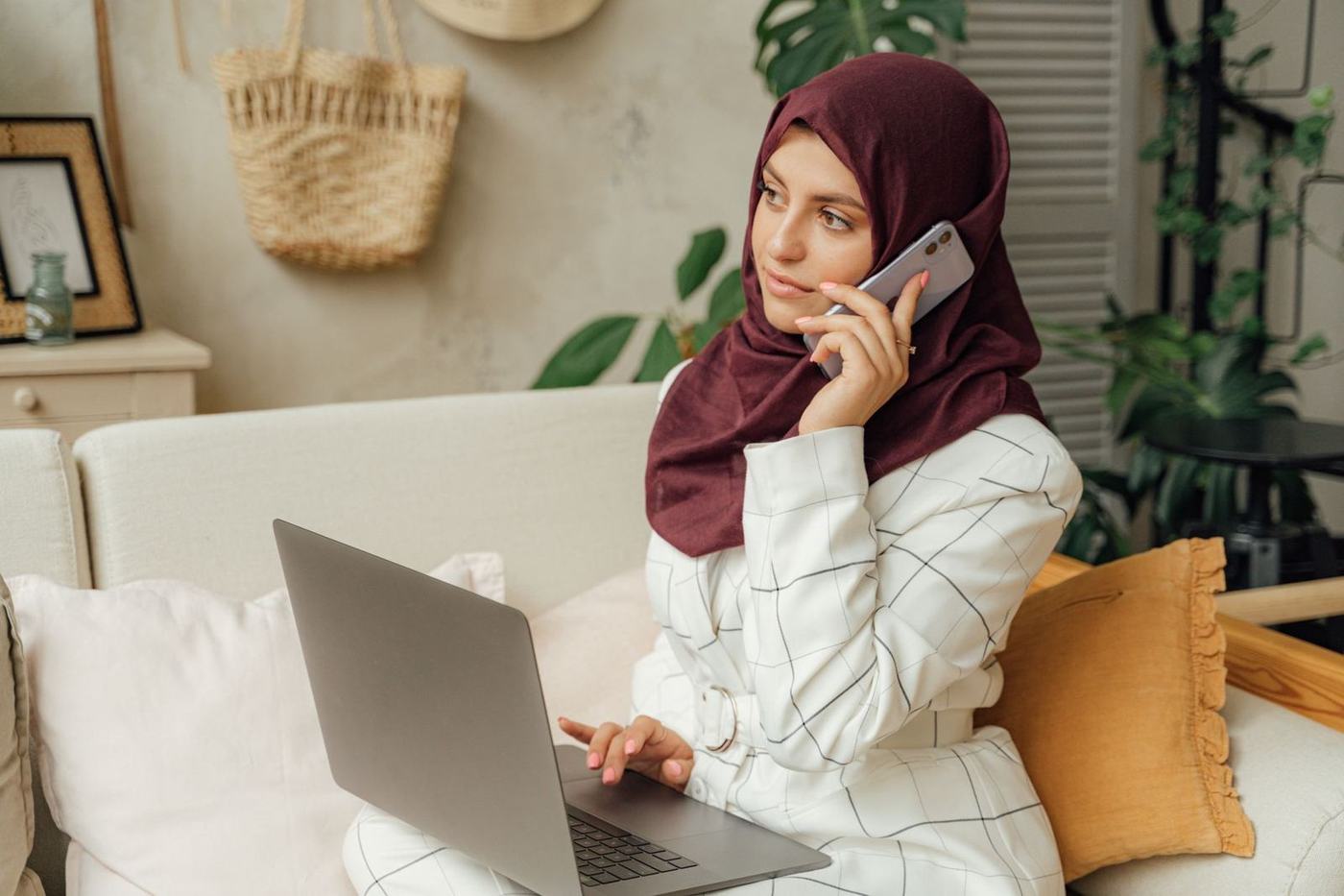امرأة بالحجاب تتحدث في الهاتف في المنزل وحاسوبها المحمول مفتوح