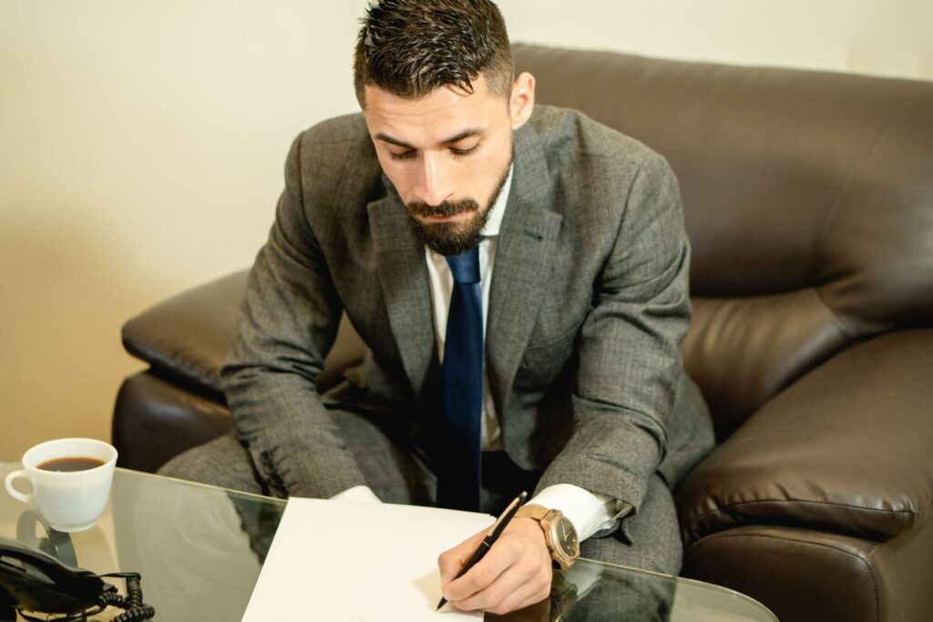 A man in a grey suit signing a paper at a glass table
