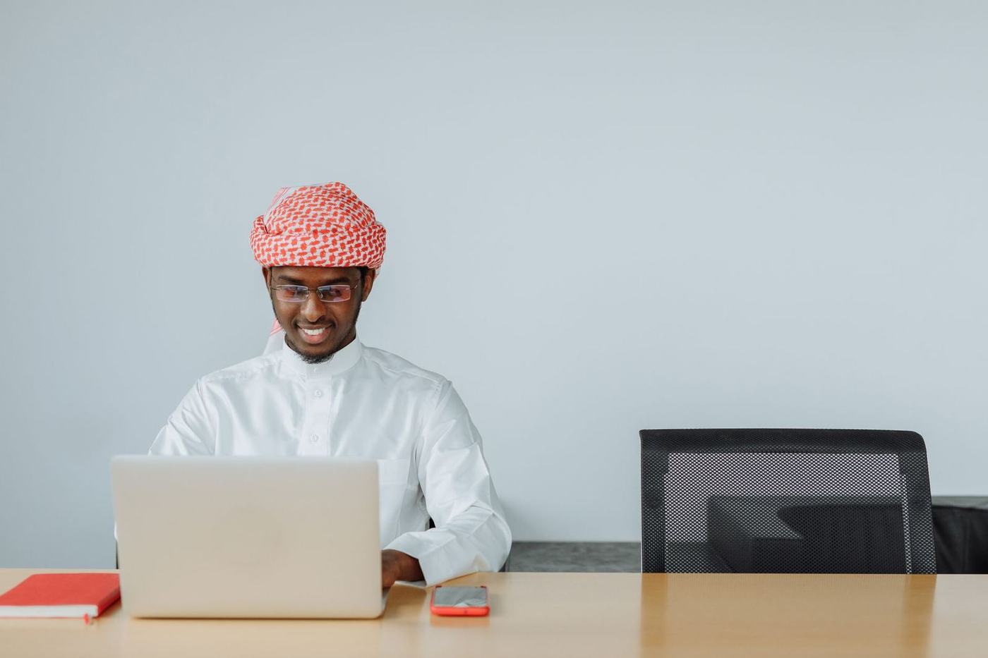 رجل بثوب أبيض وغترة حمراء يبتسم أمام حاسوبه المحمول على مكتب