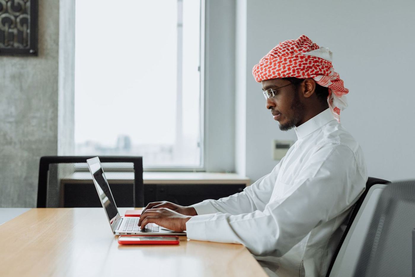 رجل بثوب أبيض وغترة حمراء يعمل على حاسوب محمول على مكتب