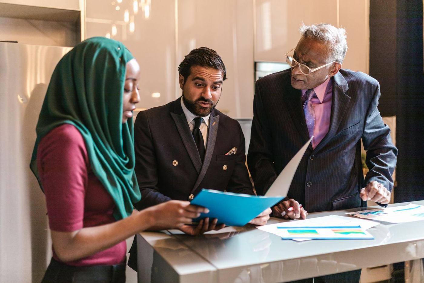 A woman in a green hijab and two men in suits going through a document in a blue folder at an office desk