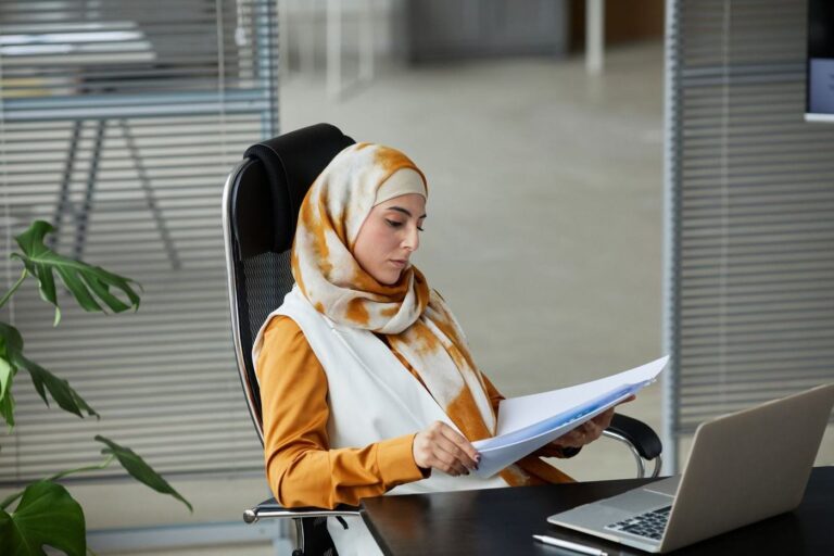 A woman in a hijab reading printed pages at her desk beside a laptop