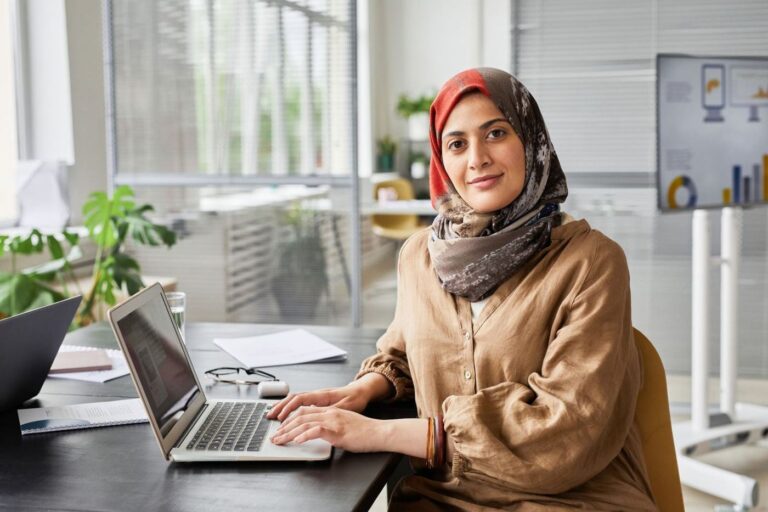 A woman in a hijab working at an office desk with a laptop and documents