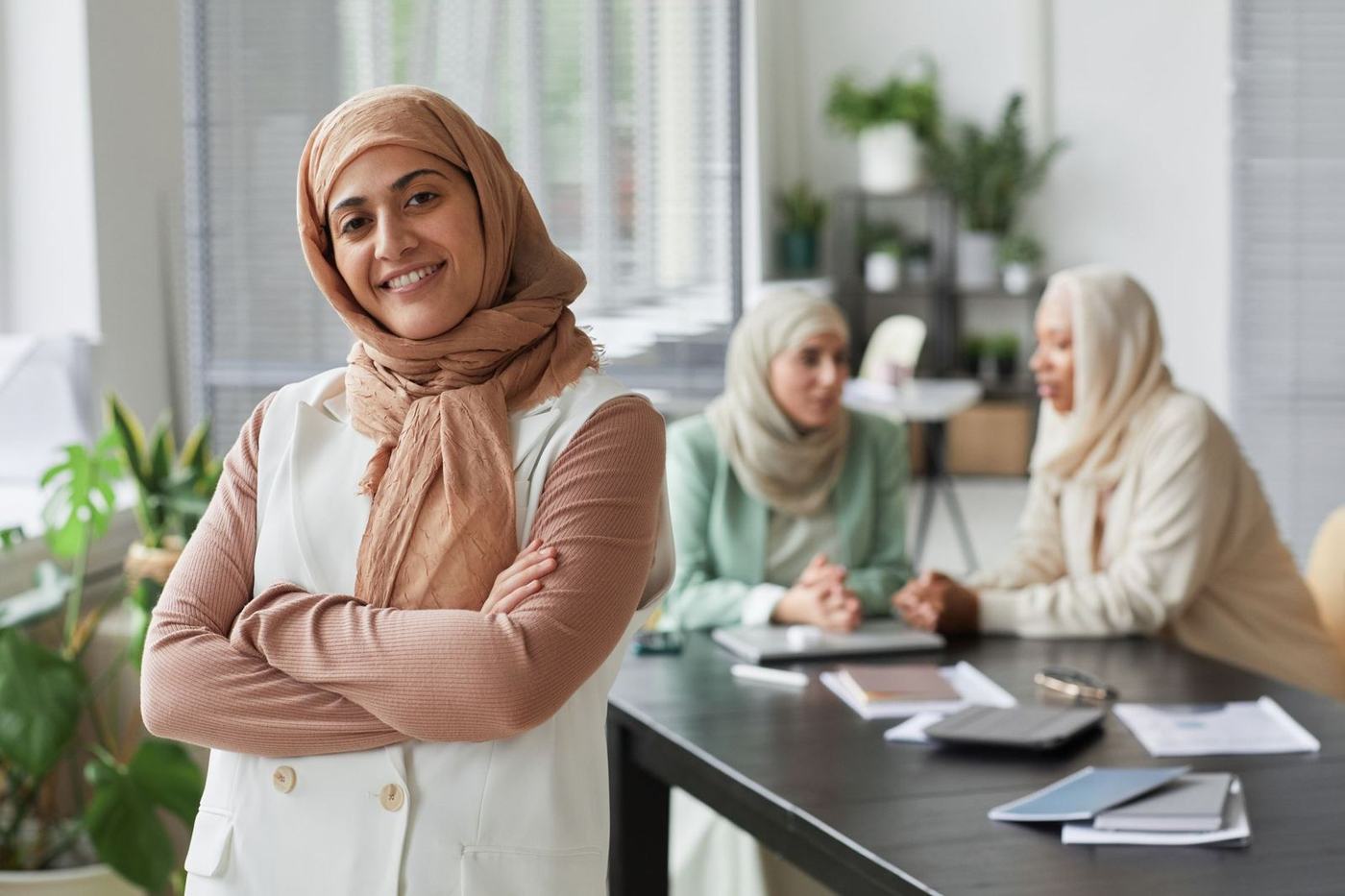 امرأة مبتسمة بالحجاب تقف في مكتب، وخلفها زميلان إلى مكتب