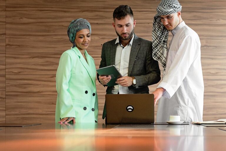Three colleagues around a laptop in an office, one man in a white thobe