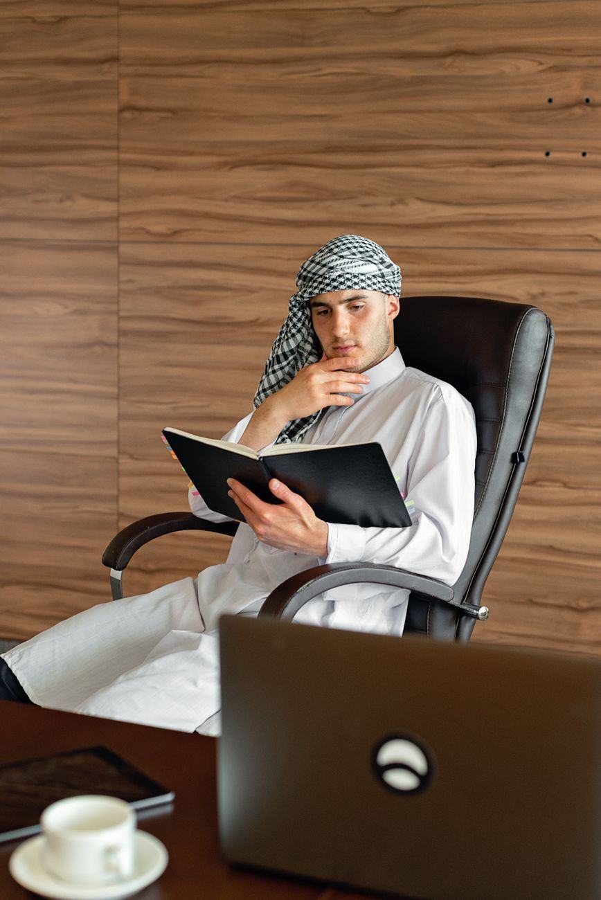 رجل بثوب أبيض وغترة مقلمة يقرأ دفترًا على مكتبه