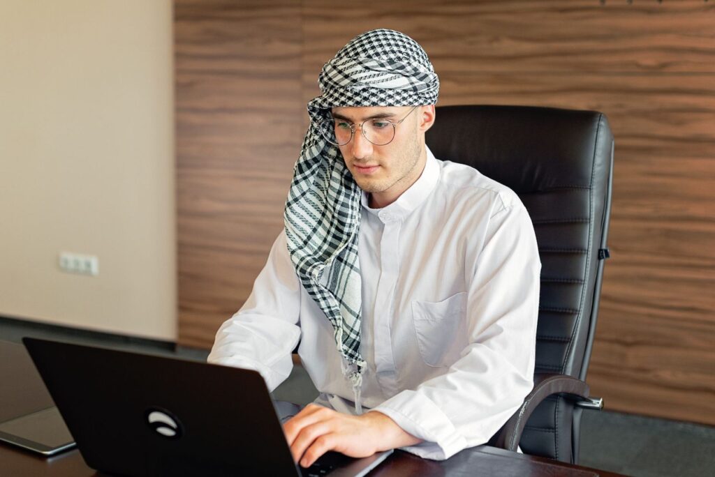 A man in a white kandura and checked headscarf working at an office desk with a laptop