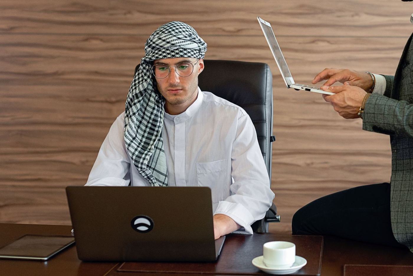 رجل بثوب أبيض وغترة مقلمة أمام حاسوب محمول بينما يناوله زميل جهازًا لوحيًا