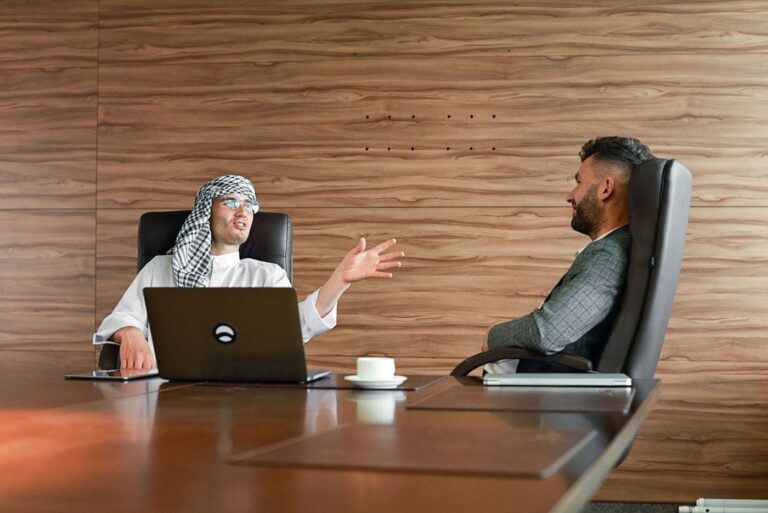 Two men talking across an office desk, one in a white thobe and headscarf
