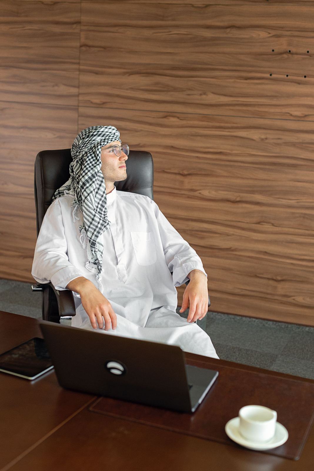 Man in a white kandura and checked ghutra seated at a boardroom desk with a laptop
