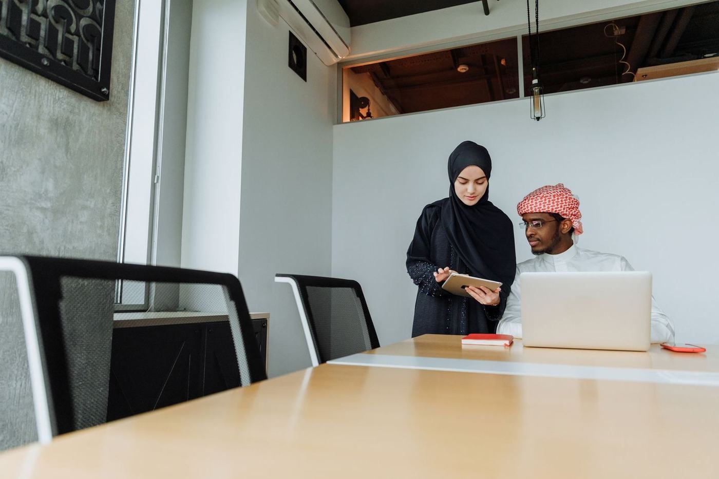 Woman in a black hijab showing a tablet to a man in a kandura at a meeting table