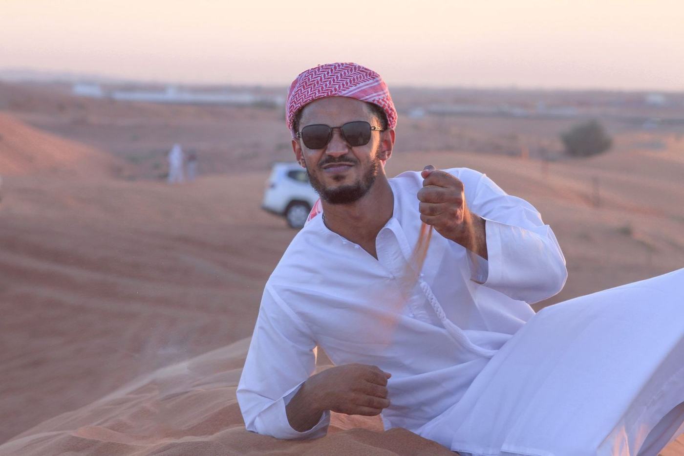 Man in a white kandura and red ghutra in the desert with a car parked behind him
